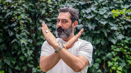 A middle-aged man with fair skin and a beard, outdoors in a wooded area, making a "no" gesture with his hands by crossing them, while looking at the camera