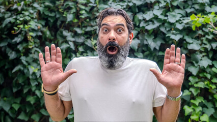 A middle-aged, fair-skinned, bearded man looking at the camera with a surprised expression in a wooded area