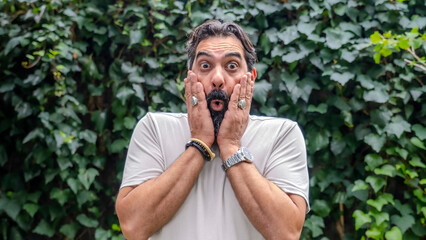 A middle-aged, fair-skinned, bearded man looking at the camera with a surprised expression in a wooded area