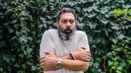 A middle-aged, fair-skinned, bearded dark-haired man looking at the camera with a facial expression showing that he's cold, set in an outdoor, wooded area