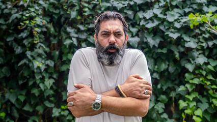 A middle-aged, fair-skinned, bearded dark-haired man looking at the camera with a facial expression showing that he's cold, set in an outdoor, wooded area
