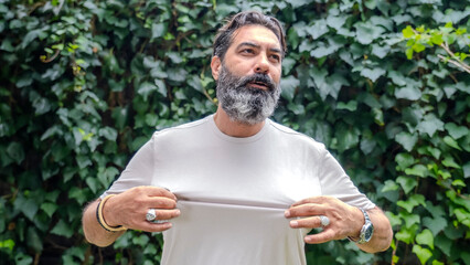 A middle-aged, dark-haired man with a beard and fair skin, outdoors in a wooded area, looking at the camera in close-up with an expression showing discomfort from the heat