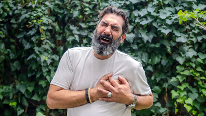 A middle-aged, white-skinned, bearded, dark-haired man in a wooded area, looking at the camera while placing his hand on his chest as if he's having a heart attack
