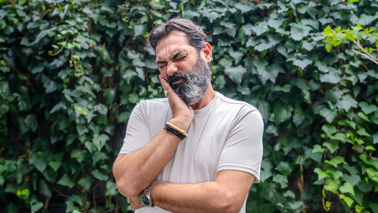 A middle-aged, fair-skinned, bearded, dark-haired man in a wooded area, holding his chin and showing signs of tooth pain while looking at the camera in a close-up shot