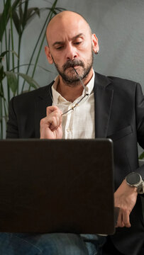 A handsome middle-aged businessman in a suit and glasses is posing outside the office environment, working on his laptop with a happy, sly, and know-it-all attitude.