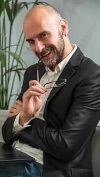 A handsome middle-aged businessman in a suit and glasses is posing outside the office environment, working on his laptop with a happy, sly, and know-it-all attitude.