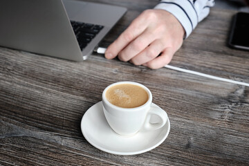 Close up of man's hand plugging laptop charging cable near white espresso cup. Conceptual image of energy gain.