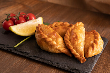 Argentine empanadas on a black slate plate with a lemon wedge, cherry tomatoes on the vine, and cilantro, placed on a wooden table.