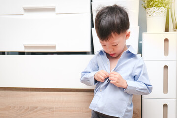 A 4-year-old Asian kindergarten boy focuses on buttoning his shirt by himself in the dressing room at home. Childhood independence, self-care skills, and early development, Soft and selective focus
