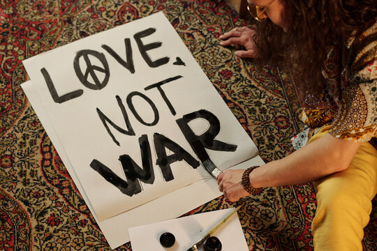 Capturing moment of person painting a peace sign and anti-war message on a large poster. Focus on handwritten message on white paper on ornate carpet flooring