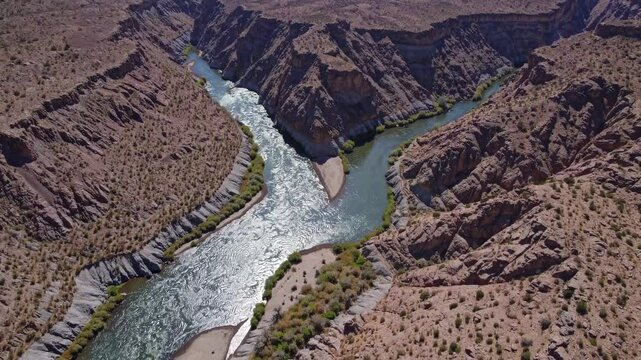 A stunning view of a river meandering through a rugged desert terrain. The rocky cliffs frame the waterway, showcasing patches of lush vegetation alongside the banks.