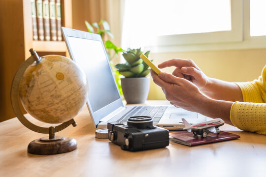 Traveler using a smartphone and laptop for booking and planning an exciting trip, surrounded by a globe, camera, airplane model, and passport on a desk - Powered by Adobe