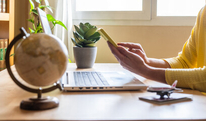 Travel agent browsing on a mobile phone and working on a laptop while booking a vacation, surrounded by a globe, airplane model, and passport on the desk