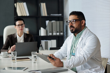 Side view portrait of mature Middle Eastern man as doctor wearing lab coat and holding clipboard while giving report during meeting in clinic office