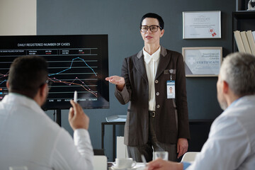 Waist up portrait of mature woman addressing audience while speaking at medical conference giving presentation standing by digital board