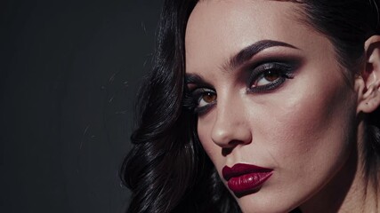 A close-up view of a woman's face showcasing stunning makeup with bold eyes and dark lipstick. The soft lighting highlights her elegant features, creating an alluring effect.