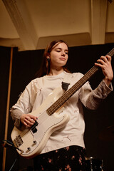 Obraz premium Vertical shot of confident teenage female artist hitting chord with deft fingers on electric bass guitar while playing punk rock music in recording studio