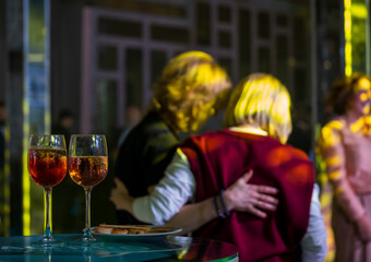 Two friends embrace during a lively party with wine glasses on table and dim lighting.