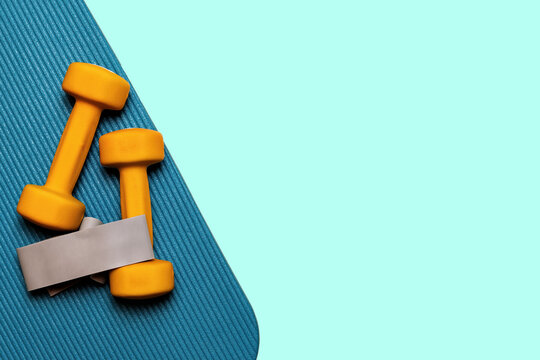 Yellow dumbbells and a resistance band rest on a teal yoga mat against a light blue background. Perfect for fitness, health, and wellness. Copy space