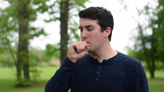 Young Caucasian man coughing outdoors, seasonal illness and respiratory health concern for flu, cold, or allergies. Sick person covering mouth, infection prevention, and healthcare awareness in nature