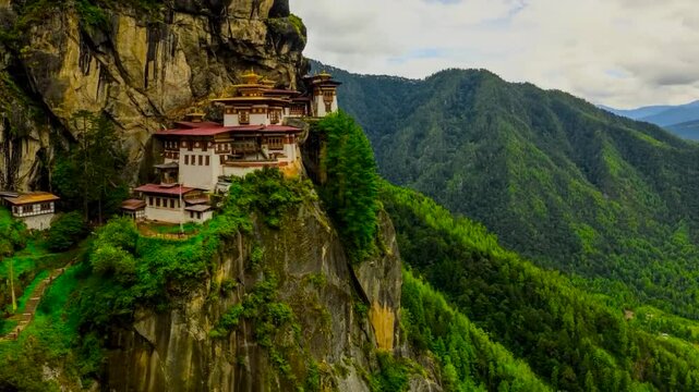 Paro Taktsang or Palphug Monastery on a steep mountain cliff during a timelapse at a sunny and cloudy day with cloud shadows running over the monastery and nature. People are seen walking in location.