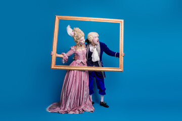 Fototapeta premium Young couple dressed in extravagant baroque costumes holding a golden frame against a vibrant blue background