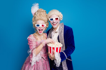 A joyful couple dressed in vintage costumes, wearing 3D glasses against a bright blue backdrop, enjoys popcorn.