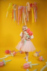 A little girl in a pink dress with balloons in the form of flowers on a yellow background.