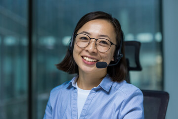 Asian businesswoman wearing a headset and eyeglasses smiles warmly at the camera in an office environment.