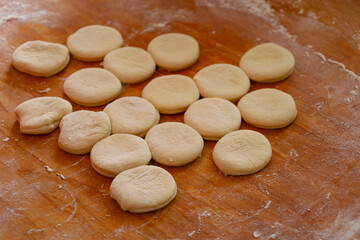 Round pieces of dough lie on a floured wooden board. Some pieces are perfectly shaped, others are slightly deformed. The dough is light, with a slight texture.