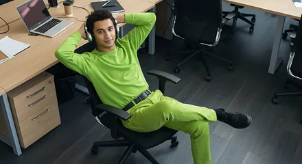 Rolgordijnen zonder boren Young man in vibrant lime green attire relaxes in his modern office chair listening to music with headphones © Tanousch