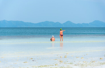 An elderly couple enjoys a relaxing beach vacation on a sunny, clear day. The husband sits on the beach in swimming trunks, while the wife, in a bikini, stands pointing to the sea in front of them.