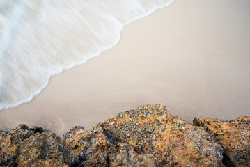 Top view of a clean brown sandy beach with rocks at the bottom and soft foaming waves crashing from above at dusk. Summer outdoor nature holiday serenity. Background, copy space or space for text.