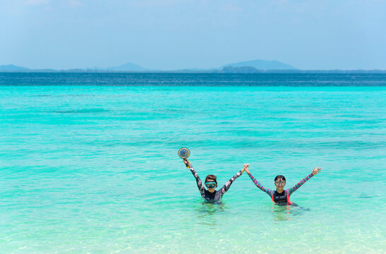 Asian mother and child tourists in long-sleeved UV-protective swimsuits and snorkeling masks enjoy playing in the clear blue water, hold hands, raise their hands to greet the camera. copy space.