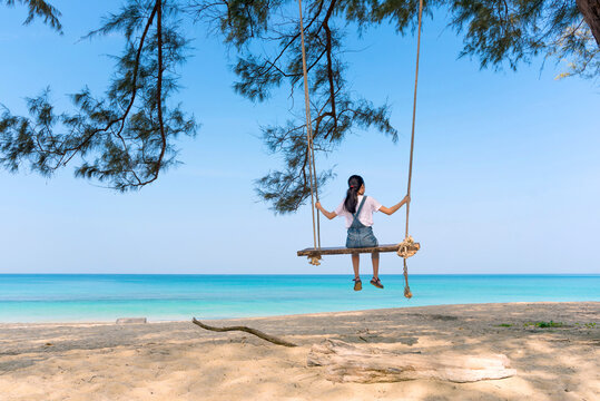 A young Asian girl sits facing the sea and plays on a swing under a pine tree by the clear blue sky and sea. A young woman is enjoying a summer day and traveling for leisure. Trang, Thailand.