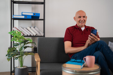 Happy retired man using mobile phone at home, sitting on comfortable sofa in living room with books and coffee cup on table, enjoying modern technology and online communication