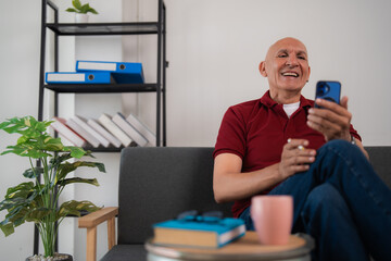 Happy retired man sitting on a comfortable sofa, holding a smartphone and smoking a cigarette, enjoying leisurely moments in the cozy living room filled with personal touches