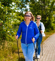 Nordic walking - two middle aged beautiful women exercising on sidewalk using Nordic walking poles...