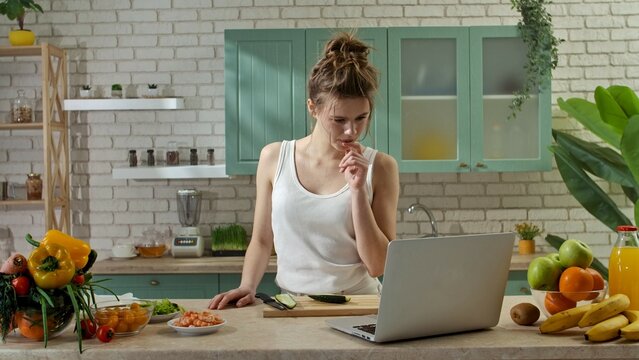 Young woman at the table in the kitchen preparing spring rolls for healthy lunch, woman eating cucumber and watching recipe video on laptop.