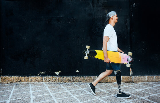 Young man with leg prosthesis holding skateboard passing a wall