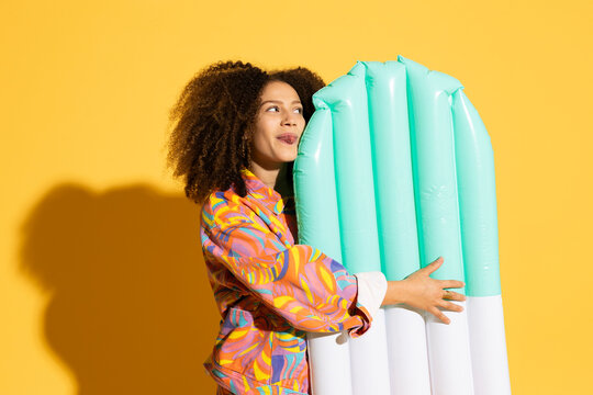 Playful young woman sticking out tongue near inflatable pool raft against yellow background
