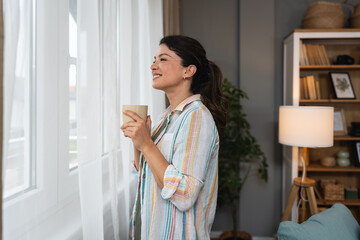 Beautiful young woman drinking a cup of coffee while looking forward standing next to the window at home. Morning routine and waking up with hot beverage. Relaxation and happiness.