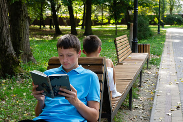 A photo captures two young boys on a bench in a sunny park. One boy engrossed in a book, while the...