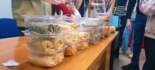 plastic containers with pancakes. People stand around, some reaching for the food.