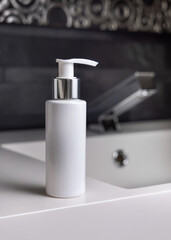 White cream bottle near modern faucet on basin in dark grey bathroom close up, cosmetic mockup