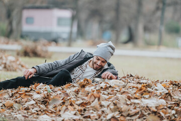 Young man smiling while lying on a pile of autumn leaves, enjoying the outdoors in a park. The scene captures the essence of fall and relaxation within a natural environment.
