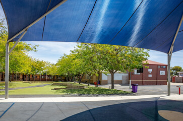 View from under a blue shade sail in a primary school campus with neatly maintained green spaces.A...