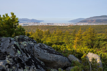 View from the mountain to the seaside town. In the foreground are rocks and thickets of Siberian dwarf pine. In the distance, on the shore of the sea bay, the city of Magadan. Magadan region, Russia.