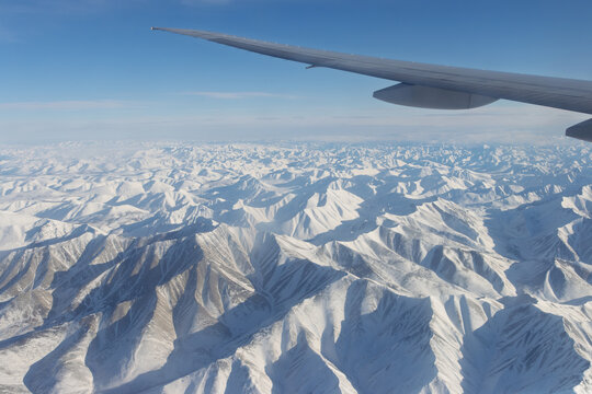 View from a flying airplane on snow-capped mountains and an airplane wing. Beautiful winter mountain landscape. Flight on a passenger plane over a mountainous area. Aviation tourism and travel.