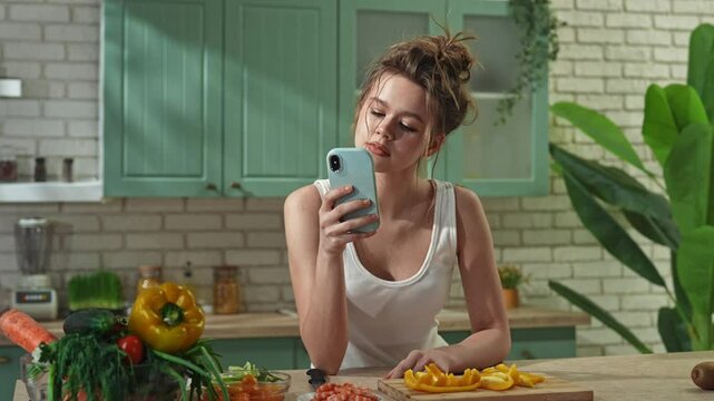 Young woman at the table in the kitchen preparing spring rolls for healthy lunch, woman texting on smartphone, thinking and eating slice of bell pepper. Slow motion.